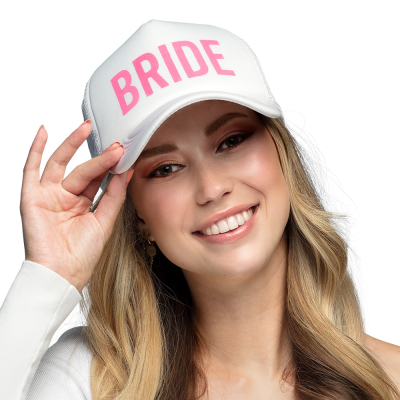 A smiling woman with long blonde wavy hair is wearing a white cap printed with 'Bride' in large pink letters.