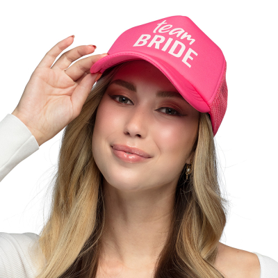 A smiling woman with long blonde wavy hair is wearing a pink cap printed with the text 'Team Bride' in large white letters.