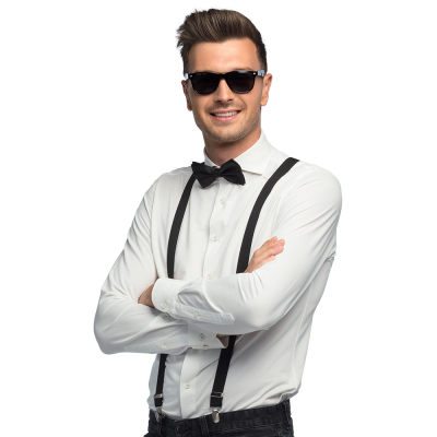 Lachende man in witte blouse met donkere spijkerbroek staat met zijn armen over elkaar en draagt een zwarte partybril, bretels en vlinderstrik, wat onderdelen zijn van een accessoireset.