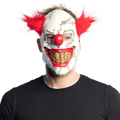 Man wears a Halloween latex mask of a broken procelain horror clown with red hair on the sides, a big red nose and a creepy big grin with yellow teeth.