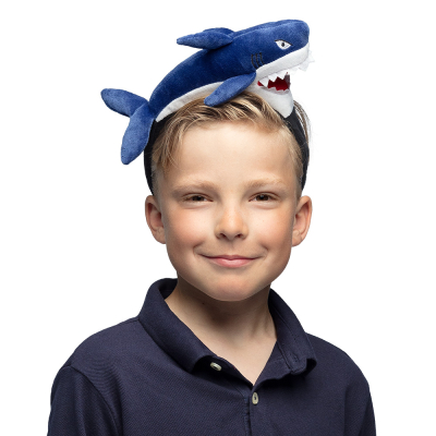 Smiling blonde boy is wearing a dark blue polo shirt and has on his head a plush tiara with a small shark on it.